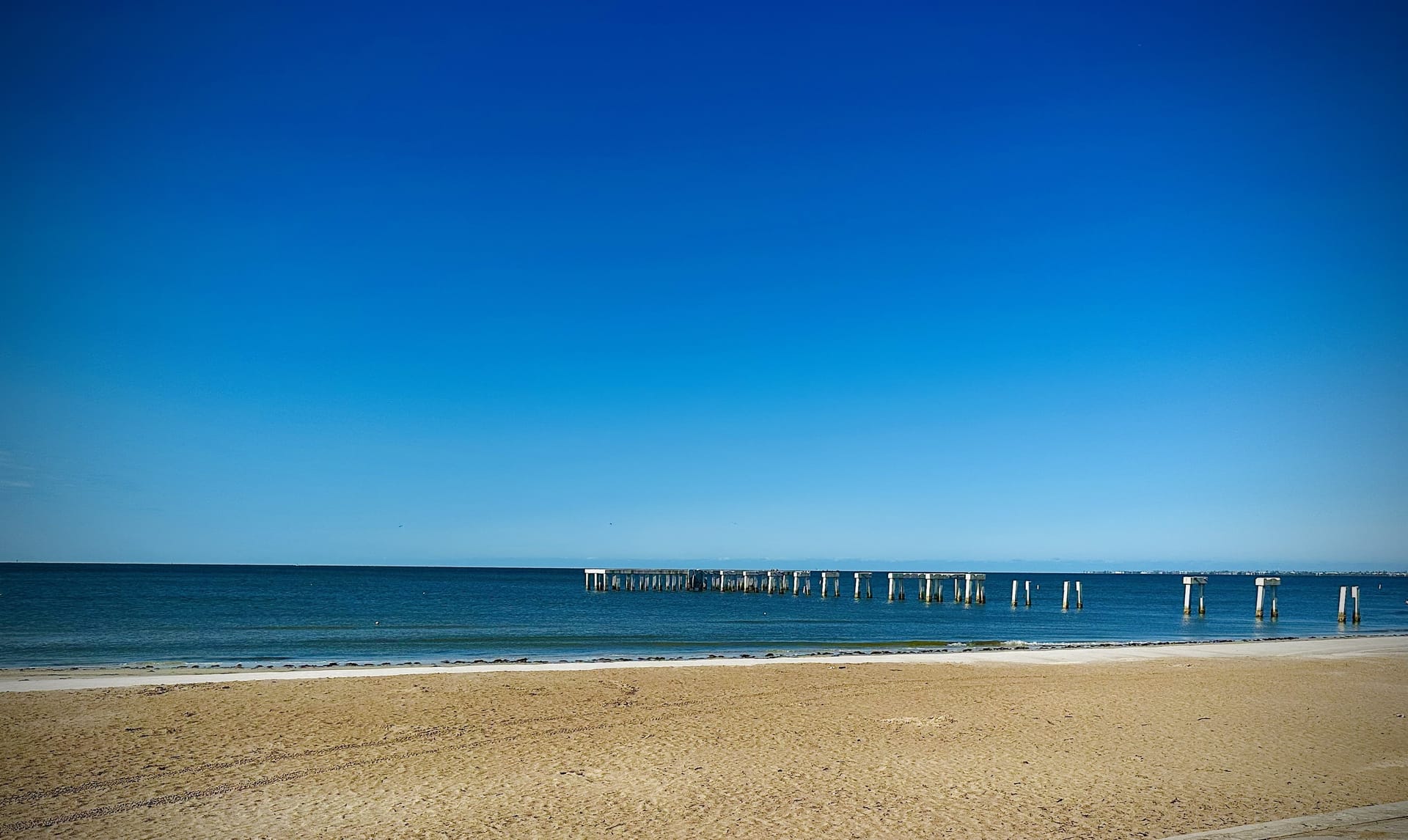 A calm beach with an old pier stretching into still blue water under a clear sky